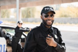 happy-african-american-police-officer-looking-at-camera-while-holding-radio-set-on-blurred-background-outdoors
